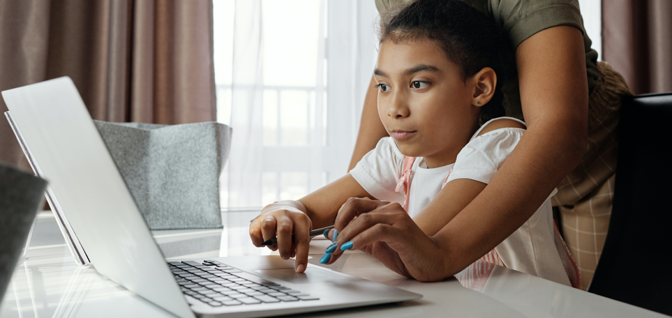 A young girl is sitting at a table in front of a laptop. Someone is reaching from behind her to help the girl use the computer.