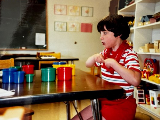 A young girl is in a classroom setting. She's seated in front of a table; there are cups on the table in many different colors.