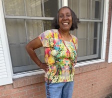 Roselyn, a woman with Down syndrome, is standing in front of a house and smiling. She's wearing a colorful blouse and jeans. Her hands are on her hips.