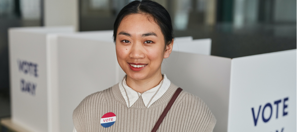 A young woman is looking at the camera and smiling. She's in a polling place and is wearing a "vote" sticker.