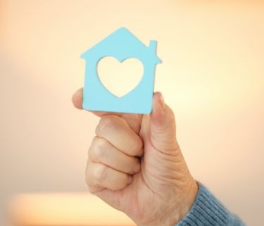A hand holds a small wooden blue house with a heart shape cut out of it.
