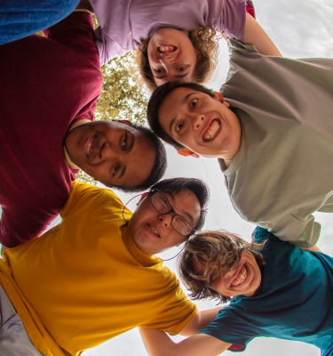A diverse group of young adults with disabilities stand together in a circle with their heads close, smiling. The view point is looking up at them from inside the circle.