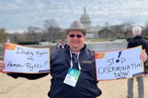 A man with disabilities stands outside. He is holding hand written signs that read, "Disability Rights are Human Rights!!" and "Stop Discrimination Now." The U.S. Capitol building is in the background.