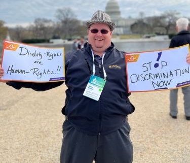 A man with disabilities stands outside. He is holding hand written signs that read, "Disability Rights are Human Rights!!" and "Stop Discrimination Now." The U.S. Capitol building is in the background.