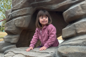 A young girl with Down syndrome is outdoors. She's standing inside a rock structure and smiling.