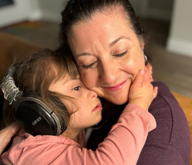 A young girl with Down syndrome is wearing headphones and hugging her mom.