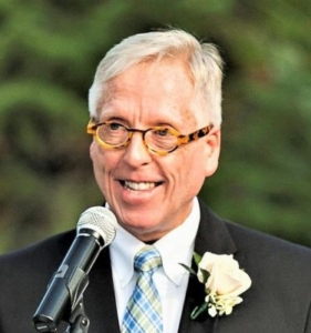 A photo of Ken Oakes wearing a suit with a flower in the lapel. He is outside at an event, speaking into a microphone.