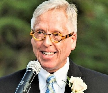 A photo of Ken Oakes wearing a suit with a flower in the lapel. He is outside at an event, speaking into a microphone.