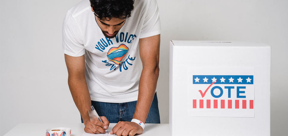 A man is leaning over a table with a white ballot box that says "Vote." He's writing something on a paper and next to him is a roll of voting stickers. He's wearing a T-shirt that says, "Your Voice. Your Vote."