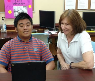 A Hispanic young man with disabilities sits with a teacher at a desk. He is looking at a laptop.