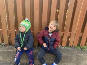 Two young boys, one with Down syndrome, wear warm, winter jackets, and are sitting together outside.