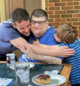 A man with WAGR syndrome sits at a table in a restaurant. He is hugging and being hugged by another man and a young boy.