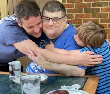 A man with WAGR syndrome sits at a table in a restaurant. He is hugging and being hugged by another man and a young boy.