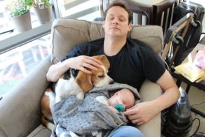 Burt and his newborn son sitting in a reclining chair, sleeping together while a dog looks at the baby.