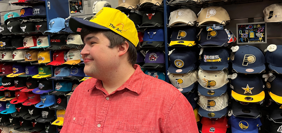 Young man displays his new hat custom embroidered with The Arc logo. In the background is a wall lined with hats, each displaying a different sports team.
